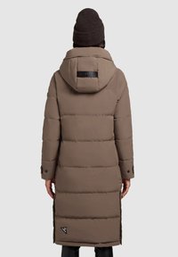 Hnedý dlhý prešívaný kabát s kapucňou, s čiernou logovou nášivkou, bočnými vreckami na zips a rukávmi s gombíkmi. Je viditeľná textúrovaná tkanina.