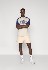 Bež sportska majica s tamnoplavim rukavima s logotipom "ADIDAS", uparena s bež kratkim hlačama. Bijele tenisice i čarape upotpunjuju odjevnu kombinaciju.