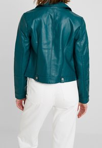 Turkusowa skórzana kurtka, dopasowany krój, ze strukturalnymi ramionami, detalami szwów i zatrzaskami u dołu. Gładka, błyszcząca tekstura.