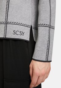 Šedý pletený svetr s černými švy a vyšitým textem "SCSY" na lemu, v kombinaci s černým oděvem.