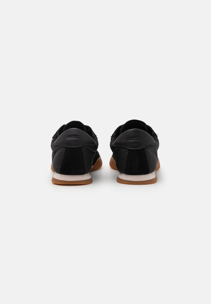 Chaussures en daim noir avec un dos en cuir lisse, dotées d'une semelle en caoutchouc beige et d'une semelle intermédiaire blanche. Logo de la marque visible à l'arrière.