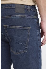 Tummansiniset farkut, joiden takataskussa on hienovaraiset ompelu-yksityiskohdat ja musta nahkainen merkkilaatta vyötäröllä.