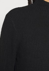 Musta ribbineulottu neulepaita, jossa on korkea kaulus ja teksturoitu kangas. Muotokieli on minimalistinen ilman näkyviä kuvioita tai yksityiskohtia.