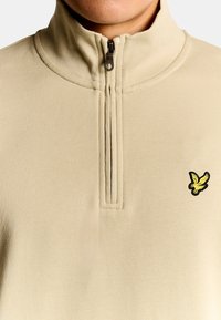 Beiger, strukturierter Pullover mit einem Reißverschluss am Kragen, mit einem kleinen gelben Vogel-Logo auf der linken Seite.