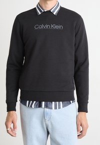 Felpa nera con colletto a girocollo e logo Calvin Klein in grigio chiaro. Presenta una camicia a righe sotto e jeans azzurri chiari.