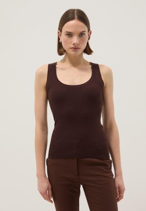 Femme aux cheveux courts et bruns porte un haut côtelé sans manches brun foncé et un pantalon marron assorti, debout devant un fond clair uni.
