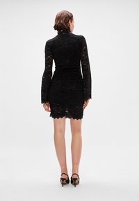 Μαύρο δαντελένιο φόρεμα με μακριά μανίκια, ψηλό λαιμό και εφαρμοστή σ silouette. Λεπτομέρειες με κυματιστό στρίφωμα, απεικονίζεται από πίσω.