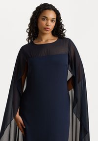 Abito blu navy fluente con maniche a capesce trasparenti, tessuto morbido, scollo rotondo e drappeggio sottile. Presenta una silhouette aderente e fessure laterali.