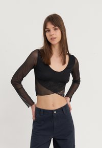 Crni kratki crop top dugih rukava s prozirnom mrežastom tkaninom i čipkastim slojem, s okruglim izrezom i prilagođenim siluetom.