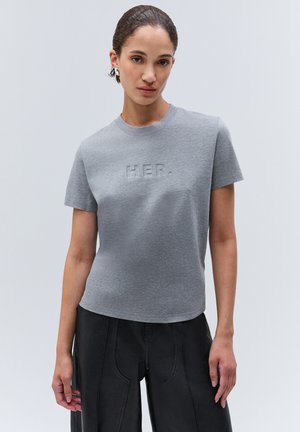 Femme aux cheveux tirés en arrière porte un t-shirt gris cintré avec le texte en relief "HER." et un pantalon noir ample sur un fond clair uni.