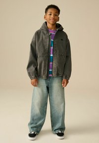 Chaqueta con capucha gris y puños elásticos, camiseta de cuello redondo morada con rayas horizontales, pantalones vaqueros anchos descoloridos y zapatillas negras.