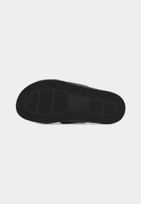 Semelle de chaussure noire avec des motifs de grip texturés et des sections de bande de roulement rectangulaires sur un fond gris clair uni.