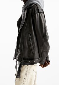 Personne portant une veste en cuir noire par-dessus un sweat à capuche gris avec un pantalon crème, se tenant devant un fond blanc.