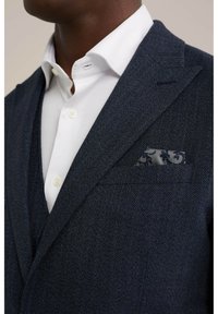 Blazer en chevrons bleu marine avec un revers pointu et une poche poitrine ornée d'un mouchoir à motif gris ; sous une chemise blanche à boutons.