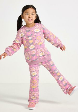 Ung pige, der balancerer på ét ben iført en lyserød blomster sweatshirt og matchende bukser med lyserøde sneakers på hvid baggrund.