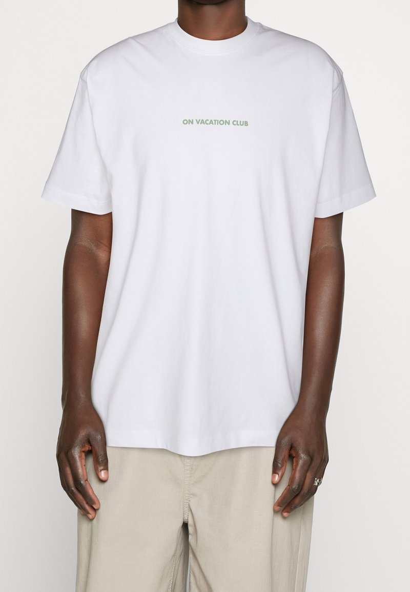 Homme portant un t-shirt blanc uni avec le texte "ON VACATION CLUB" en petite police verte sur la poitrine, associé à un pantalon beige.