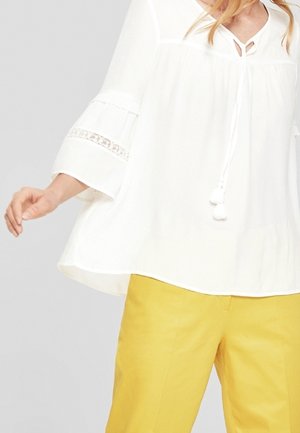 Blusa bianca, ampia, con maniche a campana, dettagli in pizzo e un nodo al collo, abbinata a pantaloni gialli.