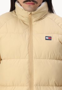 Gewatteerde jas in lichtbeige met een hoge kraag, voorzien van een ritssluiting aan de voorkant en een logo patch aan de linkerzijde. De stof lijkt zacht en geïsoleerd.