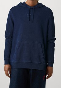 Felpa blu navy realizzata in tessuto morbido, con tasca frontale, cappuccio con lacci e logo "GAP" in rilievo sul petto. Vestibilità comoda.
