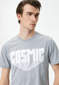Camiseta de algodón gris con un gran texto gráfico en blanco "COSMIC" con efecto 3D. Cuello redondo y mangas cortas, textura suave.