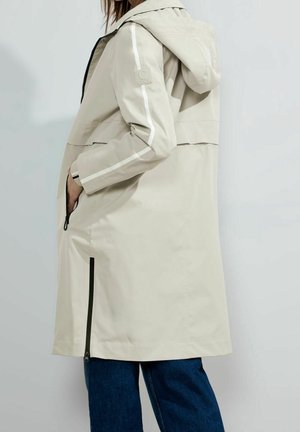 Veste imperméable beige clair avec capuche, dotée de rayures latérales blanches, de poches zippées noires et d'une coupe décontractée. Texture lisse et longueur jusqu'aux genoux.