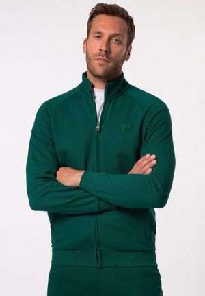 Chaqueta verde con cremallera y cuello alto, hecha de tejido suave. Presenta una cremallera frontal y un detalle de logo, con puños y bajo acanalados.