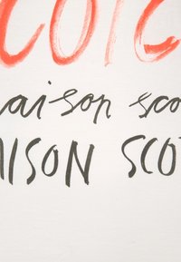 Hvidt stof med fed håndmalet skrift. Teksten inkluderer "COTON" i orange og "maison scot" i sort, med varierende tykkelse.