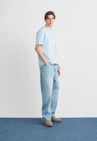 Ανοιχτό γαλάζιο βαμβακερό μπλουζάκι με τσέπη στο στήθος, σε συνδυασμό με ανοιχτόχρωμο baggy τζιν παντελόνι και μπεζ τσόκαρα, σε λευκό φόντο.