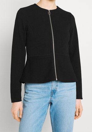 Femme portant une veste noire ajustée à manches longues avec une fermeture éclair argentée sur le devant et un jean taille haute bleu clair.