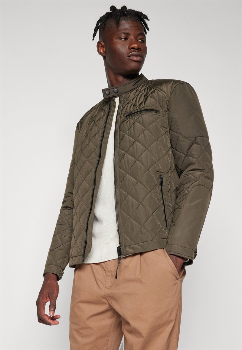 Olivgrüne gesteppte Jacke mit hohem Kragen, Reißverschluss und seitlichen Taschen. Glattes Material mit leichtem Glanz und kontrastierenden Nähten.