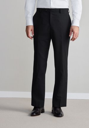 Mężczyzna w czarnych eleganckich spodniach, czarnych eleganckich butach i białej, długorękawowej koszuli zapinanej na guziki, stojący na jasnej wykładzinie podłogowej.