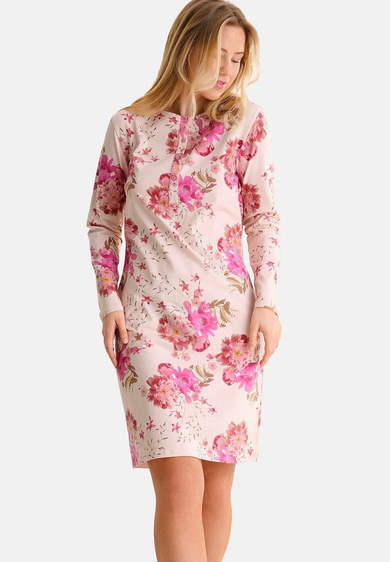 Femme portant une robe longue à manches longues de couleur rose pâle avec des motifs floraux rose vif et rouge, regardant vers le bas avec les mains reposant sur ses côtés.