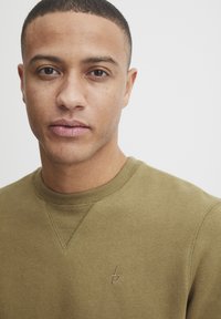 Olive green sweatshirt with a ribbed neckline and subtle embroidered logo on the chest, made from a soft, textured fabric.