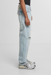 Jeans de mezclilla de corte relajado en azul claro, con un acabado desgastado, roturas en las rodillas y detalles de costura en contraste. Combinados con zapatillas grises y plateadas.
