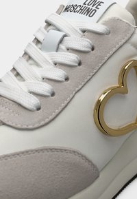 Λευκό αθλητικό παπούτσι με μπεζ σουέτ λεπτομέρειες. Διαθέτει επίπεδα λευκά κορδόνια, λογότυπο σε σχήμα χρυσής καρδιάς, υφασμένους επιφάνειες και λεία σόλα.