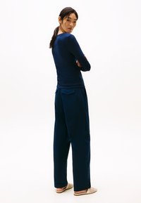 Top din material bleumarin cu mânecă lungă și siluetă ajustată, purtat cu pantaloni lejerași bleumarin, cu buzunare laterale, și pantofi slip-on din material țesut, de culoare deschisă.