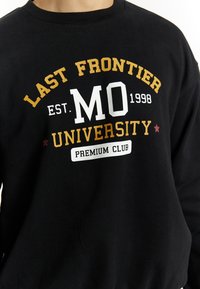 Felpa nera con testo stampato in oro, bianco e rosso. Il testo recita "LAST FRONTIER MO 1998 UNIVERSITY PREMIUM CLUB."
