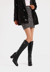 Stivali neri in pelle al ginocchio con tacco a blocco e dettagli in fibbia argentata, abbinati a un blazer nero con paillettes e una gonna grigia testurizzata.