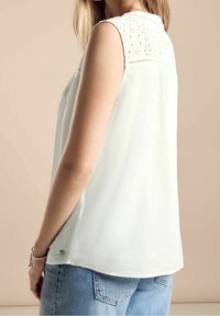 Vit ärmlös blus med spetsdetaljer på axlarna, lös passform och rundad nederkant; matchad med ljusblå denimjeans.