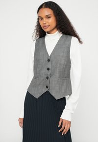 Γκρι γιλέκο με σχέδιο ψαροκόκκαλο, γιακά με εγκοπές, πέντε μαύρα κουμπιά και πλαϊνές τσέπες, φορεμένο πάνω από λευκό ζιβάγκο και μαύρη πλισέ φούστα.