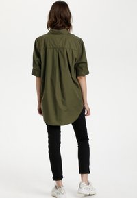 Camicia oversize verde oliva con maniche corte, orlo curvo e dettaglio sul retro, abbinata a pantaloni neri aderenti e sneaker bianche.