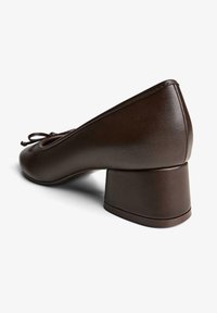 Pantof maro din piele cu toc gros, vârf rotund și detaliu decorativ cu fundă pe partea superioară. Prezintă o textură fină și un design robust.