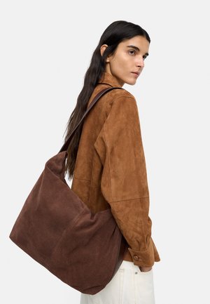 Kobieta z długimi ciemnymi włosami nosi brązową zamszową kurtkę i niesie dużą ciemnobrązową zamszową torbę na ramię.