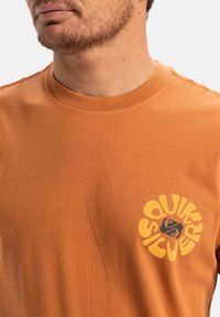 Oranžové bavlněné tričko s kulatým výstřihem. Na levé části hrudníku se nachází kruhová žlutá grafika s textem a logem.