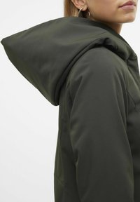 Chaqueta de softshell verde oliva, con una gran capucha estructurada y una textura suave, cuenta con bolsillos laterales y un diseño minimalista.