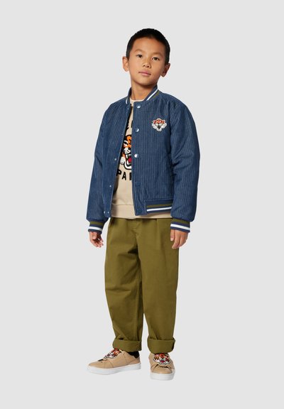 Ragazzo che indossa una giacca bomber in corduroy blu, una felpa beige con stampa tigre, pantaloni verde oliva e sneakers beige con design a tigre, in piedi.