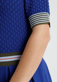 T-shirt en tricot texturé bleu avec des poignets côtelés verts et blancs rayés. Il présente un corsage ajusté et une jupe évasée.