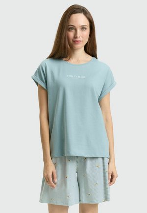 Vrouw draagt lichtblauw T-shirt met opgerolde mouwen met de tekst "Tom Tailor" en gestreepte korte broek met kleine gele bloemmotieven, staand tegen een effen achtergrond.