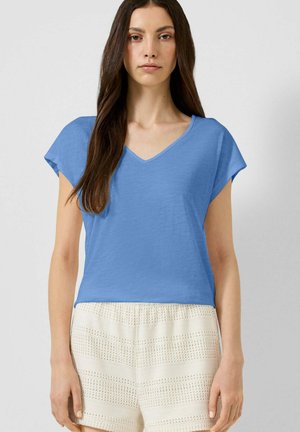 Frau mit langen braunen Haaren, die ein hellblaues V-Ausschnitt-Kurzarmtop und cremefarbene, strukturierte Shorts vor einfarbigem Hintergrund trägt.