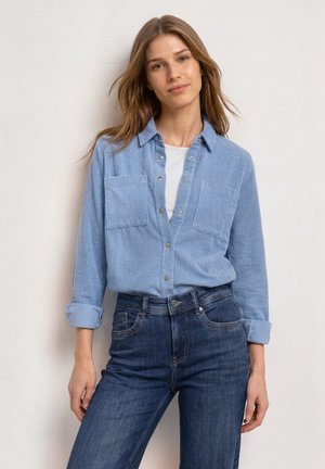 Ung kvinde med lyst brunt hår iført en lyseblå kordfløjls skjorte over en hvid top og mørkeblå jeans, står op ad en hvid væg.
